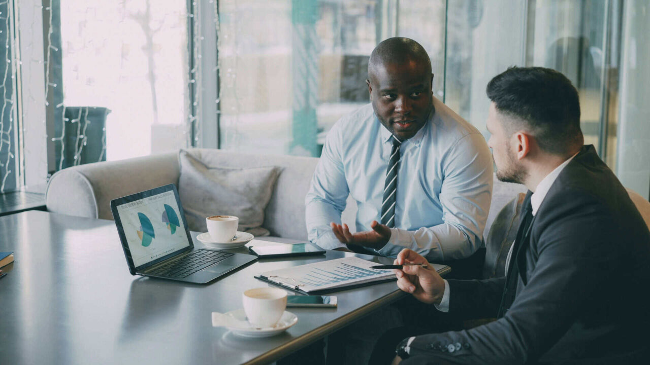 Two businessmen discussing janitorial supplies and facility solutions with charts open on a laptop.