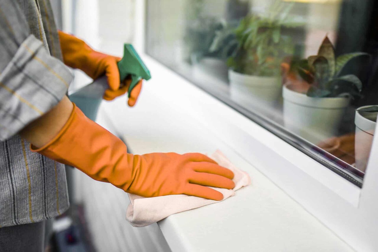 Person cleaning a window sill with a cloth and spray bottle, showcasing janitorial supplies for facility solutions.