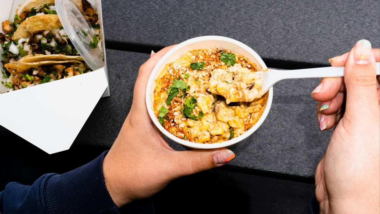 Hands holding elote corn cup with cheese and spices, next to a box of tacos on a dark table, highlighting food service supplies.