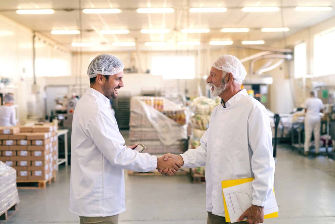 Two men in a food service facility, wearing hairnets and lab coats, shake hands, symbolizing trusted local network.