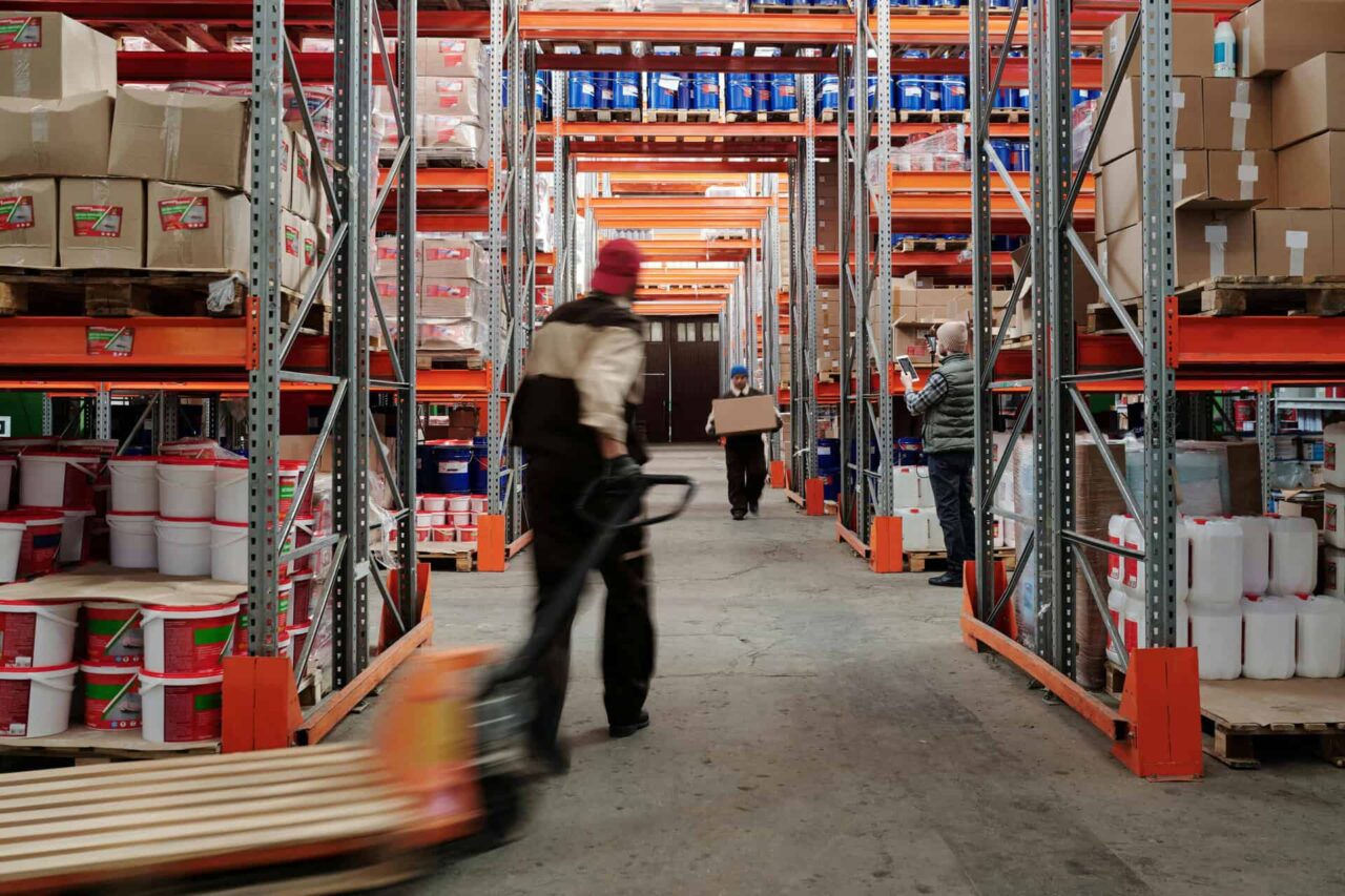 Warehouse aisle filled with stacked janitorial supplies, workers handling boxes; manufacturers representative setting.